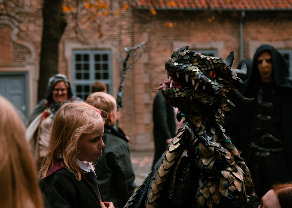 A young girl dressed as a Hogwart student observing the animated dragon hand puppet, with a mand dressed as Vordmort in the background.