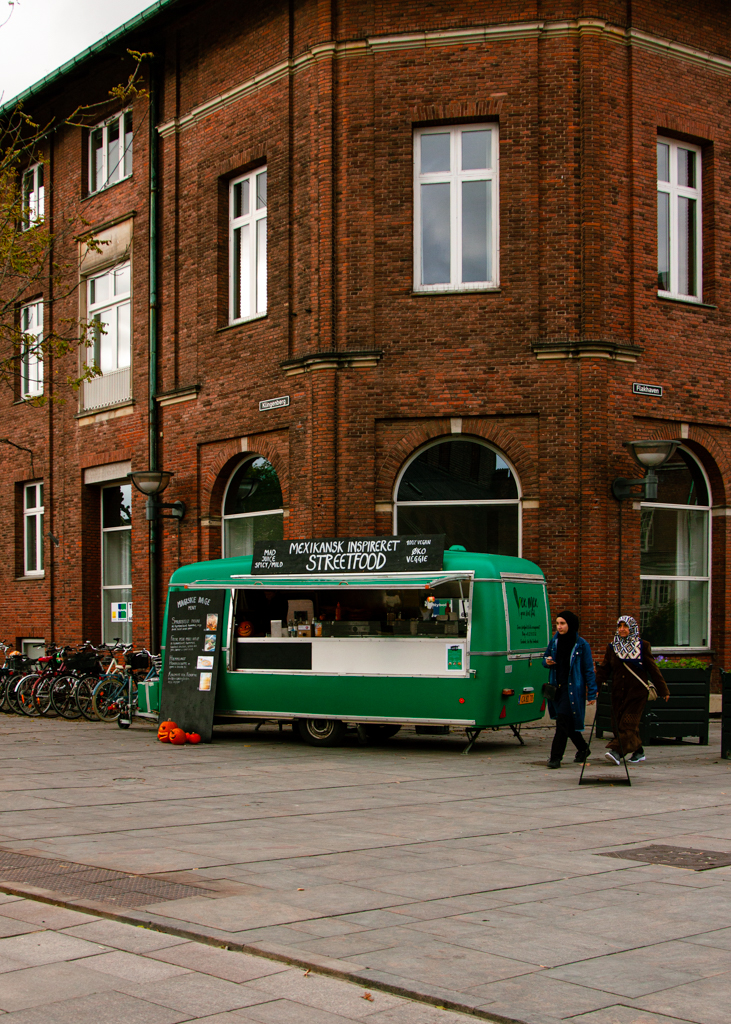 A green vintage van converted to a Mexican-inspired food truck parked on a pedatrian pathway. Two young women in hijab walking by the food truck.