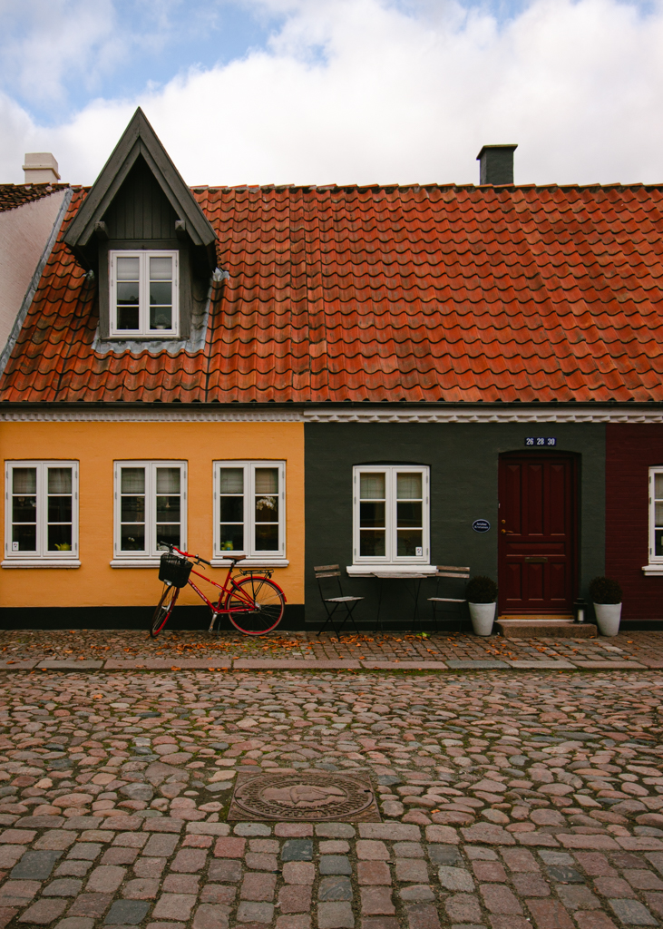 A line of two houses painted yellow and green with white framed windows, copper shingled roof and a red bike parked in front of the yellow wall. 