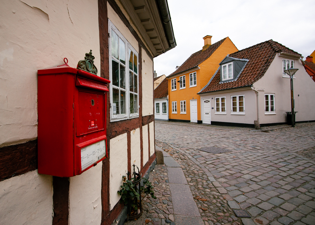 A close up photo of the red post box attached to a light beigh wall along a cobble stone pathway lined with colourful houses.