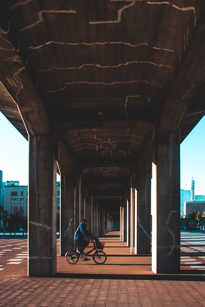 A person in a blue coat cycling toward right under an overpass bridge.