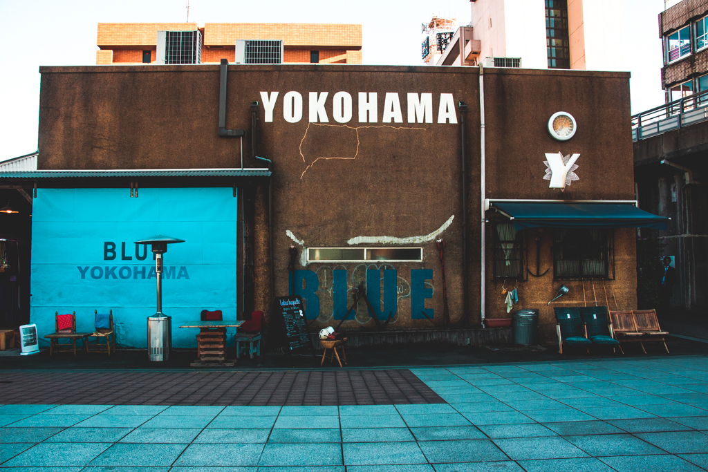 The exteror of a building with the words YOKOHAMA BLUE painted on it. In front of it, there are some folding chairs, an outdoor warmer, and an A-Frame menu.