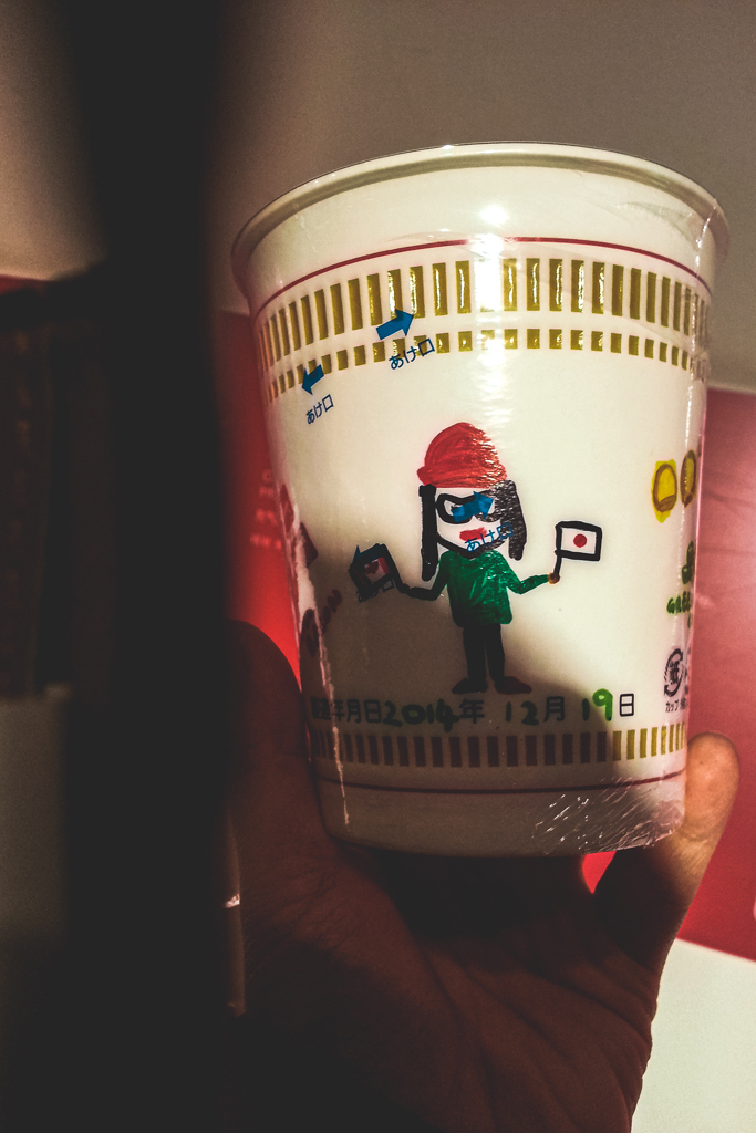 A personalized Cup Noodle by Phoebe. It is a self portrait in a green sweater, black pants and red beanie hold a Canadian flag in the right hand and a Japanese flag in the left. Below the drawing is the date in Japanese, Year 2014, Month 12 and Day 19.